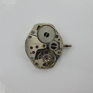 May include: A silver watch movement with a silver face and a black inscription that reads "17 Jewels".