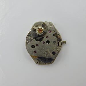 Könnte beinhalten: Ein silbernes Uhrwerk mit einem goldfarbenen Aufzugstift und mehreren kleinen Zahnrädern. Das Uhrwerk ist für eine Armbanduhr ausgelegt.