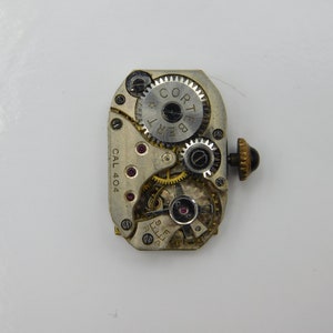 May include: Close-up of a silver watch movement with the text "Cortebert" and "Cal 404" visible. The movement has a gold-colored winding stem with a black knob.