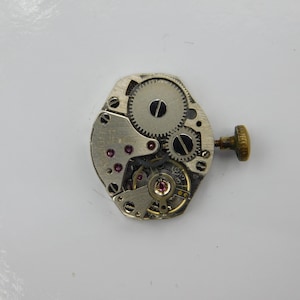 May include: A silver-toned watch movement with a winding stem and visible gears. The movement is designed for a wristwatch.