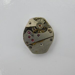 May include: A silver-toned watch movement with visible gears and red jewels. The movement is in a hexagonal shape.