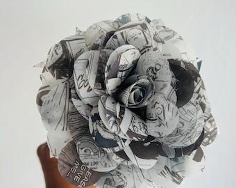 May include: A handcrafted paper flower constructed from black and white manga pages. The petals are shaped into a layered, circular bloom, featuring various printed illustrations and the visible text "PLEASE GIVE ME..." on one of the lower petals.