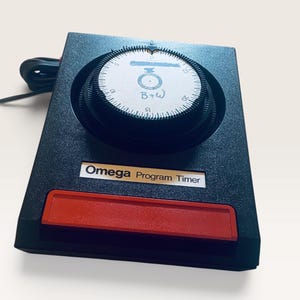 May include: A black Omega Program Timer with a large circular dial featuring white markings and numbers from 0 to 25. The device has a rectangular red button at the bottom and a black power cord extending from the side. The text B+W is written in blue ink on the center of the dial.