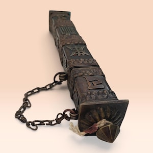 May include: A dark bronze-toned rectangular incense holder featuring intricate embossed symbols and decorative patterns on its sides. A metal chain is attached to the side, and a pointed cap with a small white cord is at the end.