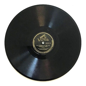 Könnte beinhalten: Eine schwarze, kreisförmige Vinyl-Schallplatte mit einem zentralen Etikett, das goldenen Text auf schwarzem Hintergrund aufweist. Das Etikett zeigt das Victor-Logo, den Songtitel In a Church in an Old Hawaiian Town und den Künstler Al Kealoha Perry and his Singing Surfriders.