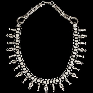 May include: A silver necklace with a detailed design. The necklace features a series of small, round, and teardrop-shaped elements connected by a delicate chain. The clasp is a simple hook and eye closure. The necklace is displayed against a black background.
