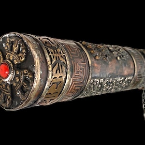 May include: A cylindrical metal case with an aged finish, featuring intricate embossed patterns and a geometric border. The circular end cap displays a raised relief design centered with a bright red stone, and a small metal chain hangs from the side.