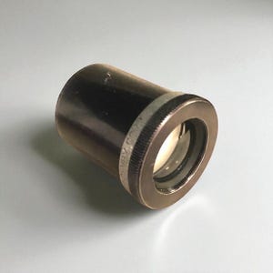 May include: A vintage brass and black telescope eyepiece. The eyepiece has a threaded ring and a lens. The words "BURY CO." are visible on the ring. The eyepiece is designed to be attached to a telescope.