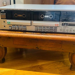 May include: A silver JVC stereo double cassette deck featuring two cassette compartments labeled A and B, a power button, a headphone jack, multiple control buttons, and a digital display panel with input level indicators on the right side.