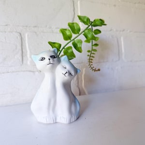 May include: A white ceramic vase shaped like two cats, with light blue accents on the ears. The cats have black facial features and are holding a green leafy plant. The vase is on a white surface.