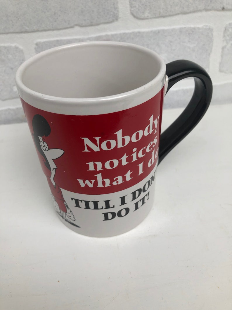Op de afbeelding: Witte keramische mok met een rode band met een stripfiguur en de tekst "Nobody notices what I do till I do it!". De mok heeft een zwart handvat en een wit interieur.