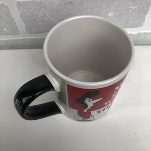 Op de afbeelding: Witte keramische mok met een zwarte handgreep en een rode vierkante afbeelding. De afbeelding toont een stripfiguur en tekst. De mok is leeg en staat op een wit oppervlak.
