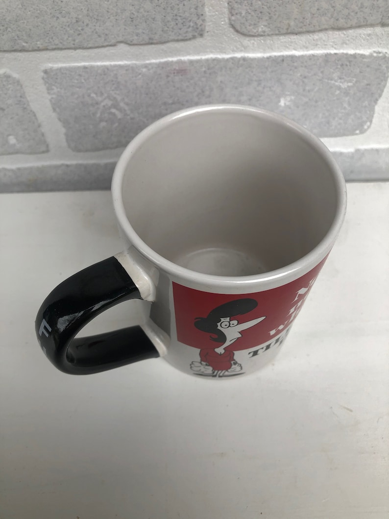 Op de afbeelding: Witte keramische mok met een zwarte handgreep en een rode vierkante afbeelding. De afbeelding toont een stripfiguur en tekst. De mok is leeg en staat op een wit oppervlak.