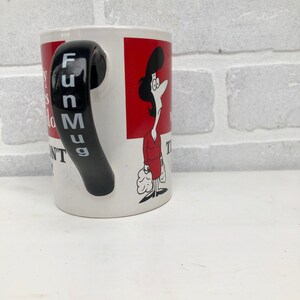 Op de afbeelding: Witte keramische mok met een zwarte handgreep met verticaal de tekst "Fun Mug". De mok heeft een cartoonillustratie van een vrouw in een rode jurk en een rode achtergrond. De mok heeft tekst.