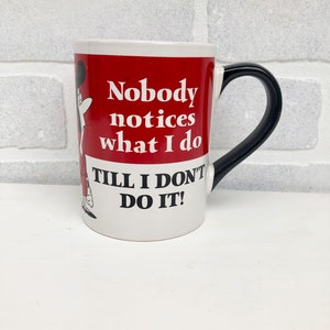 Op de afbeelding: Witte keramische mok met een zwart handvat en een rode band met de tekst "Nobody notices what I do Till I don't do it!". Een stripfiguur staat op de zijkant van de mok.