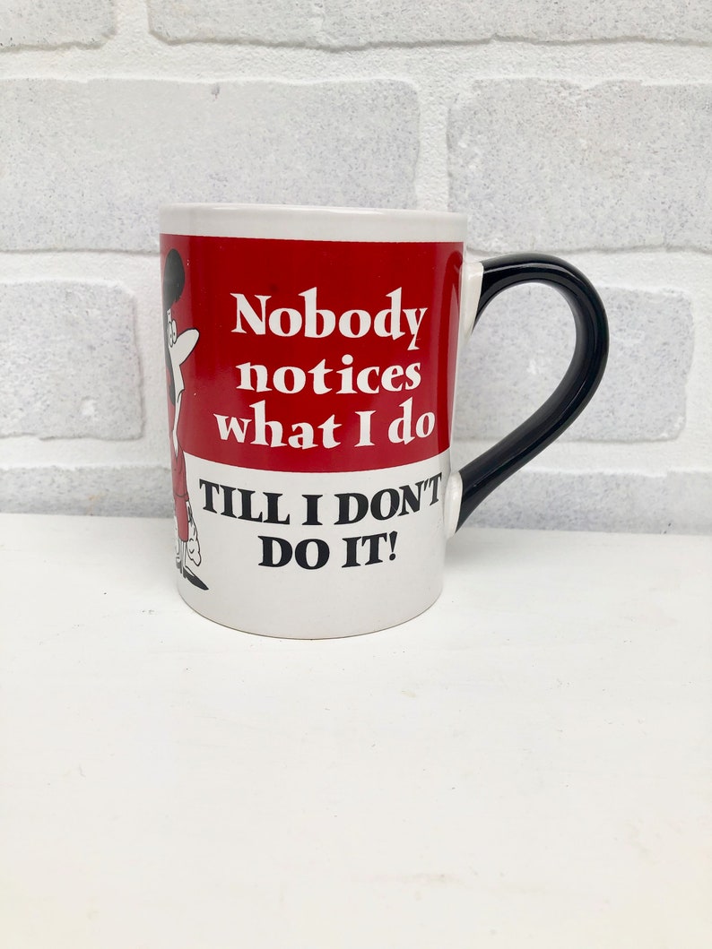 Op de afbeelding: Witte keramische mok met een zwart handvat en een rode band met de tekst "Nobody notices what I do Till I don't do it!". Een stripfiguur staat op de zijkant van de mok.