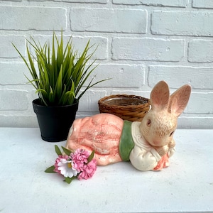 May include: A ceramic bunny figurine in a pink and white dress with green accents. The bunny is lying on its side with its head resting on its paws. The figurine is on a white surface with a green plant in a black pot in the background.