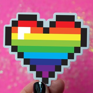 May include: A rainbow pixel heart sticker with a white outline. The sticker is on a pink background with iridescent glitter.
