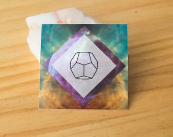 Puede incluir: Una pegatina cuadrada con un fondo de galaxia colorido. Una forma de diamante blanco con un contorno negro contiene una forma geométrica en el centro. La pegatina tiene un borde morado.