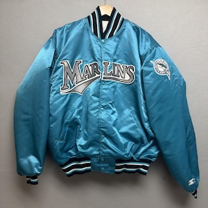 May include: A light blue satin jacket with the word "MARLINS" in large letters across the chest. The jacket has a black, white, and blue striped collar, cuffs, and hem. A team logo is on the sleeve.