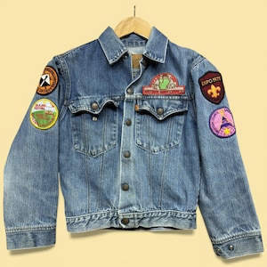 May include: A vintage denim jacket with multiple patches. The jacket is a light blue wash with a button-up front, two chest pockets, and various patches including "EXPO 1975" and "Western Area Council". The jacket is hanging on a wooden hanger.