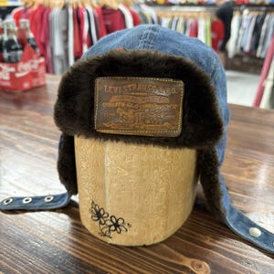 May include: A blue denim trapper hat with brown faux fur lining and ear flaps. The hat features a brown leather patch with the Levi Strauss & Co. logo. The hat is displayed on a wooden form.