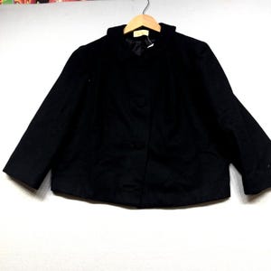 May include: A black cropped jacket featuring a rounded collar, long sleeves, and a front button closure. The garment is displayed on a wooden hanger, showcasing a simple design with a smooth texture and a visible interior lining at the neckline.