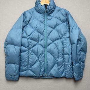 May include: A light blue puffer jacket featuring a high collar, a full-length front zipper, and a distinct diamond-shaped quilted pattern throughout the body. The jacket includes two side pockets with zipper closures and long sleeves with cuffs.
