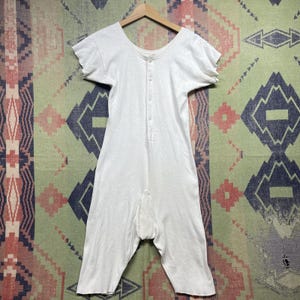 May include: A white one-piece union suit featuring short sleeves with raw edges, a scoop neckline, and a vertical front placket with four buttons. The garment is constructed from a textured knit fabric and extends to mid-thigh length with raw hems.