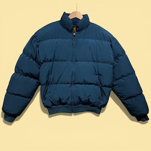 May include: A teal puffer jacket with a high collar and a full-length zipper. The jacket has a small logo on the chest and is hanging on a wooden hanger. The jacket has two side pockets and ribbed cuffs and hem.