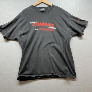 May include: A charcoal gray t-shirt with the text "Being a COMPAQ customer is a DirectPlus" in red and white. The shirt is hanging on a wooden hanger.