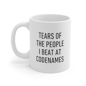 May include: White ceramic coffee mug with black text that reads "Tears of the people I beat at Codenames".