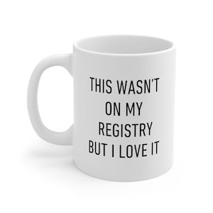 May include: White ceramic coffee mug with black text that reads "This wasn't on my registry but I love it."