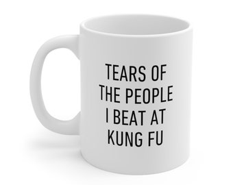 Puede incluir: Taza de cerámica blanca con asa curva. La taza presenta texto negro que dice "TEARS OF THE PEOPLE I BEAT AT KUNG FU". La taza está diseñada para bebidas calientes.