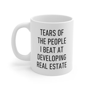 May include: White ceramic coffee mug with black text that reads "Tears of the people I beat at developing real estate."