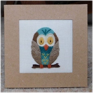 以下が含まれることがあります： 刺繍されたフクロウのアート作品。フクロウは、茶色、ティール、黄色の糸で白い背景に描かれ、正方形の薄茶色の段ボールフレームに収められています。フクロウは茶色の枝にとまっています。