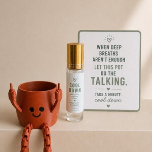 May include: A terracotta-colored pot with arms and legs, a smiling face, and a middle finger gesture. Next to it is a clear glass roller bottle with a gold cap, labeled "COOL DOWN." A card reads "When deep breaths aren't enough, let this pot do the talking."