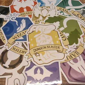 Op de afbeelding: Een verzameling kleurrijke stickers met verschillende fantasy-thema's. De stickers bevatten illustraties en tekst, waaronder "Dungeon Master", dobbelstenen en namen van klassen zoals "Wizard" en "Barbarian".