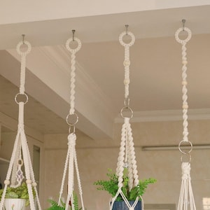 May include: Four off-white macrame plant hangers suspended from the ceiling. Each hanger features intricate knotted cord patterns, metal rings, and wooden beads. Two hangers hold blue ceramic pots with green plants, one holds a white pot, and one holds a wooden base with a white pot.