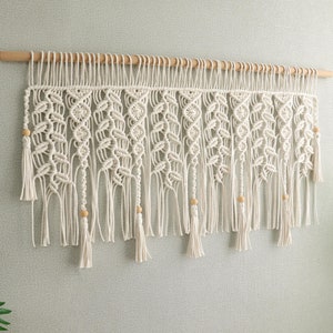 以下が含まれることがあります： 上部に木製の棒が付いた白いマクラメの壁掛け。壁掛けは、結び目とタッセルの繰り返しパターンを特徴としています。壁掛け全体に小さな木製のビーズが散りばめられています。