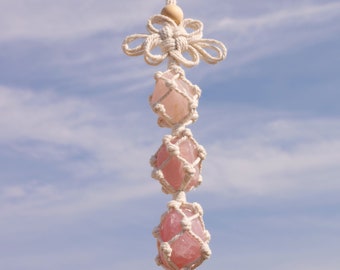 Puede incluir: Una decoración colgante de macramé con tres piedras de cuarzo rosa rosadas, envueltas en una red tejida. La parte superior tiene un nudo decorativo y una cuenta de madera, suspendidos de un cordón blanco. El fondo es un cielo azul.