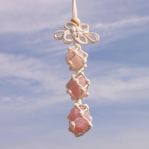 May include: A hanging macrame crystal decoration featuring three pink rose quartz stones encased in a woven net. The top has a decorative knot and wooden bead, suspended from a white cord. The background is a blue sky.