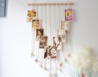 Könnte beinhalten: Ein weißer Makramee-Wandbehang mit einer Holzleiste oben. Der Makramee ist mit kleinen Holzperlen besetzt und hat Vintage-Fotos, die mit Mini-Wäscheklammern daran befestigt sind.