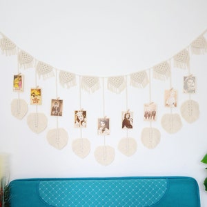 Könnte beinhalten: Eine weiße Makramee-Girlande mit Quasten hängt an einer weißen Wand. Die Girlande ist mit kleinen Fotos verziert, die mit Wäscheklammern befestigt sind. Die Girlande hängt über einem blaugrünen Sofa.