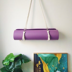 以下が含まれることがあります： 白い壁に白いロープとフックを使って紫色のヨガマットが掛けられています。マットは巻き上げられており、表面に凹凸があります。