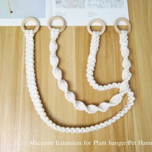以下が含まれることがあります： 木製のリングが付いた白いマクラメのプラントハンガーエクステンションが4つ。エクステンションは長さが異なり、U字型に配置されています。