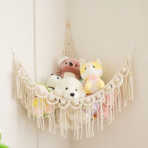 May include: A white macrame hammock with fringe hanging in a corner with stuffed animals inside. The hammock is made of cotton rope and has a natural, bohemian style.