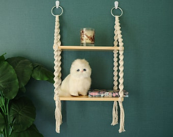 May include: A white fluffy cat sits on a wooden shelf with a candle and a book. The shelf is suspended from the wall by two macrame ropes.