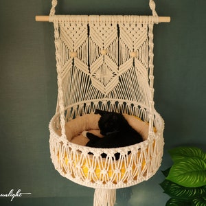 May include: A white macrame cat hammock with a yellow cushion and a black cat sleeping inside. The hammock is hanging from a wooden dowel rod and is against a green wall.