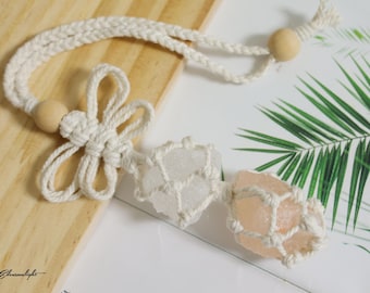 Puede incluir: Un portacristales de macramé hecho a mano con dos cristales en bruto. El soporte está hecho de cordón de algodón blanquecino y presenta un diseño en forma de flor. Los cristales son de color rosa claro y transparente. El soporte está sobre una superficie de madera.